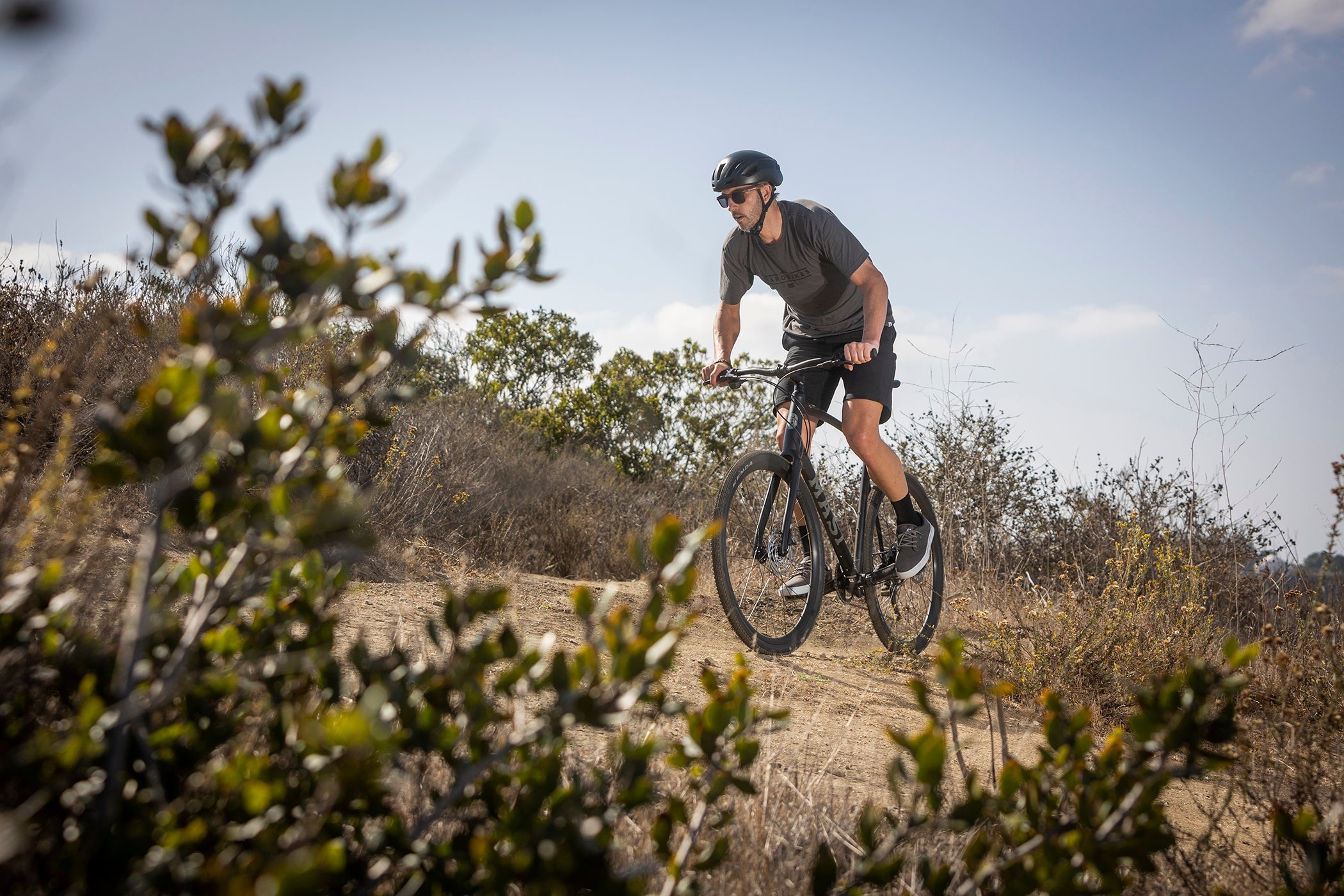 A person wearing a helmet rides a bicycle on a dirt path through a dry, shrub-filled landscape under a clear blue sky.