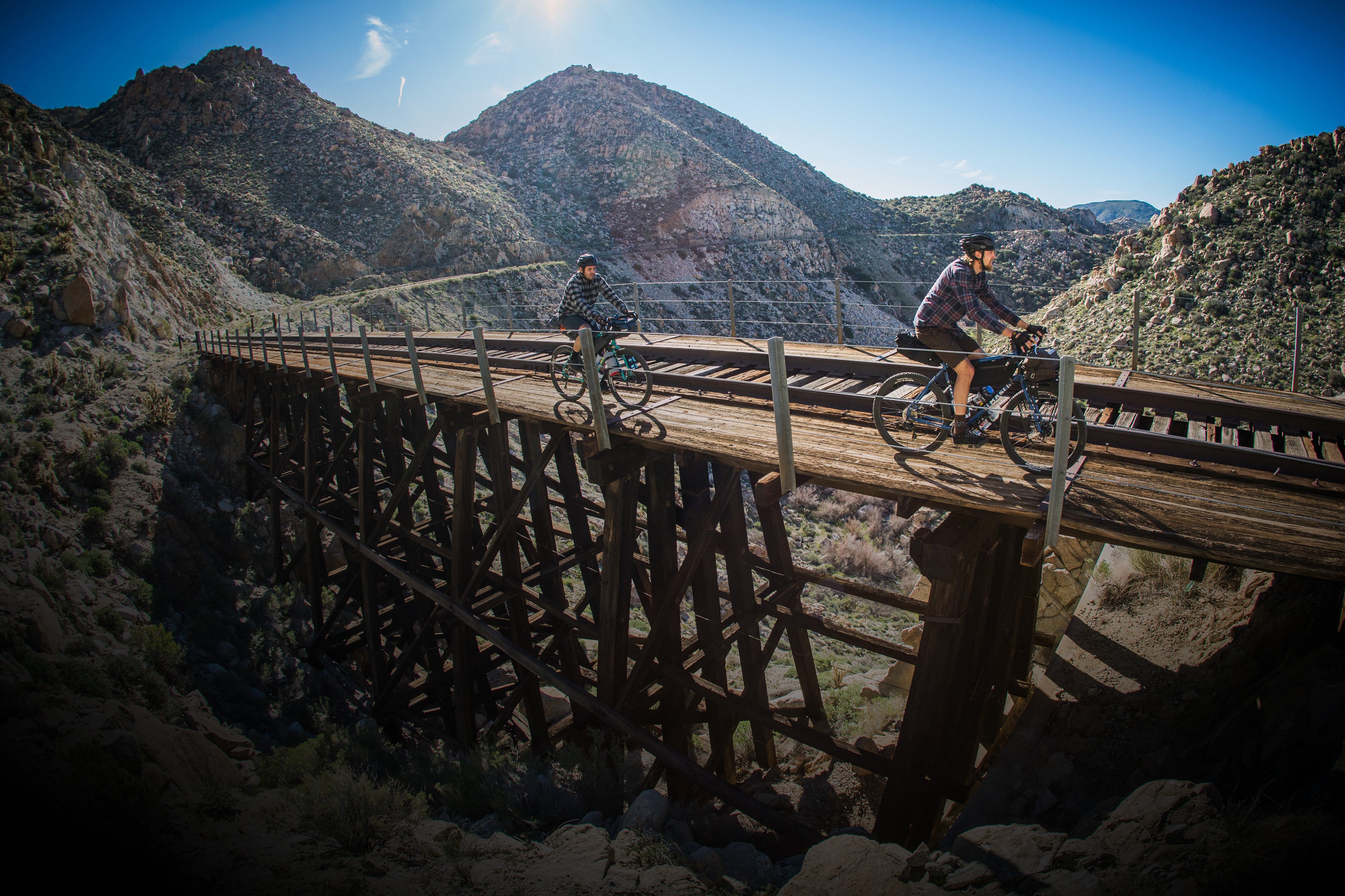 Two people ride bicycles across an old wooden trestle bridge in a mountainous area under a clear blue sky. Sunlight highlights the rugged landscape as they cycle side by side.