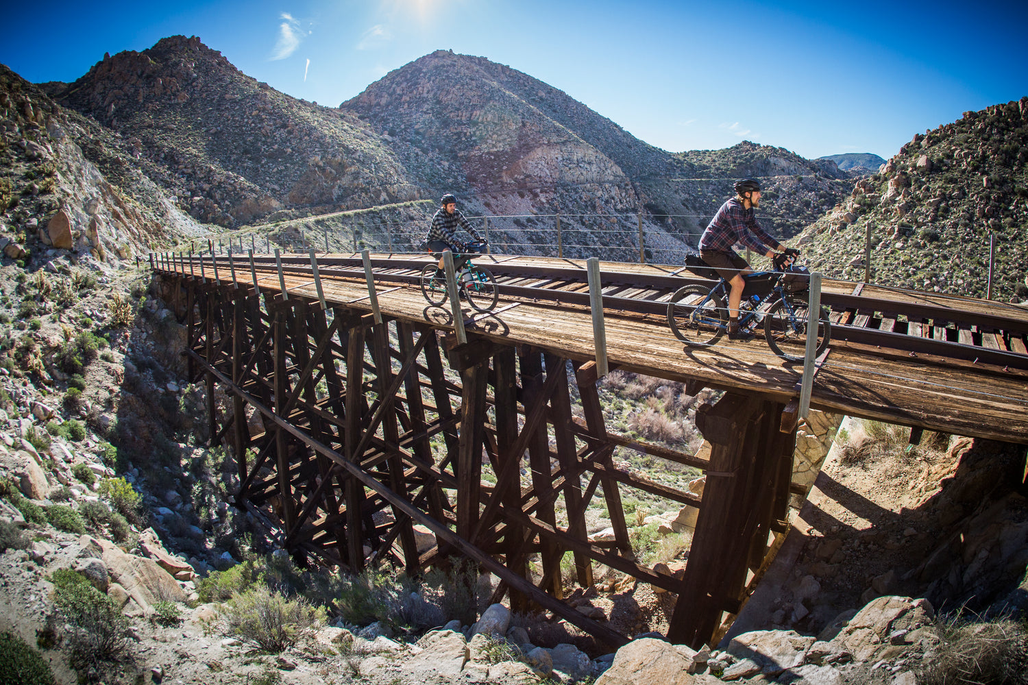 Two people ride bicycles across a wooden railway bridge, set against a backdrop of rugged mountains and a clear blue sky. The sun casts long shadows, highlighting the structure of the bridge and the surrounding rocky terrain.