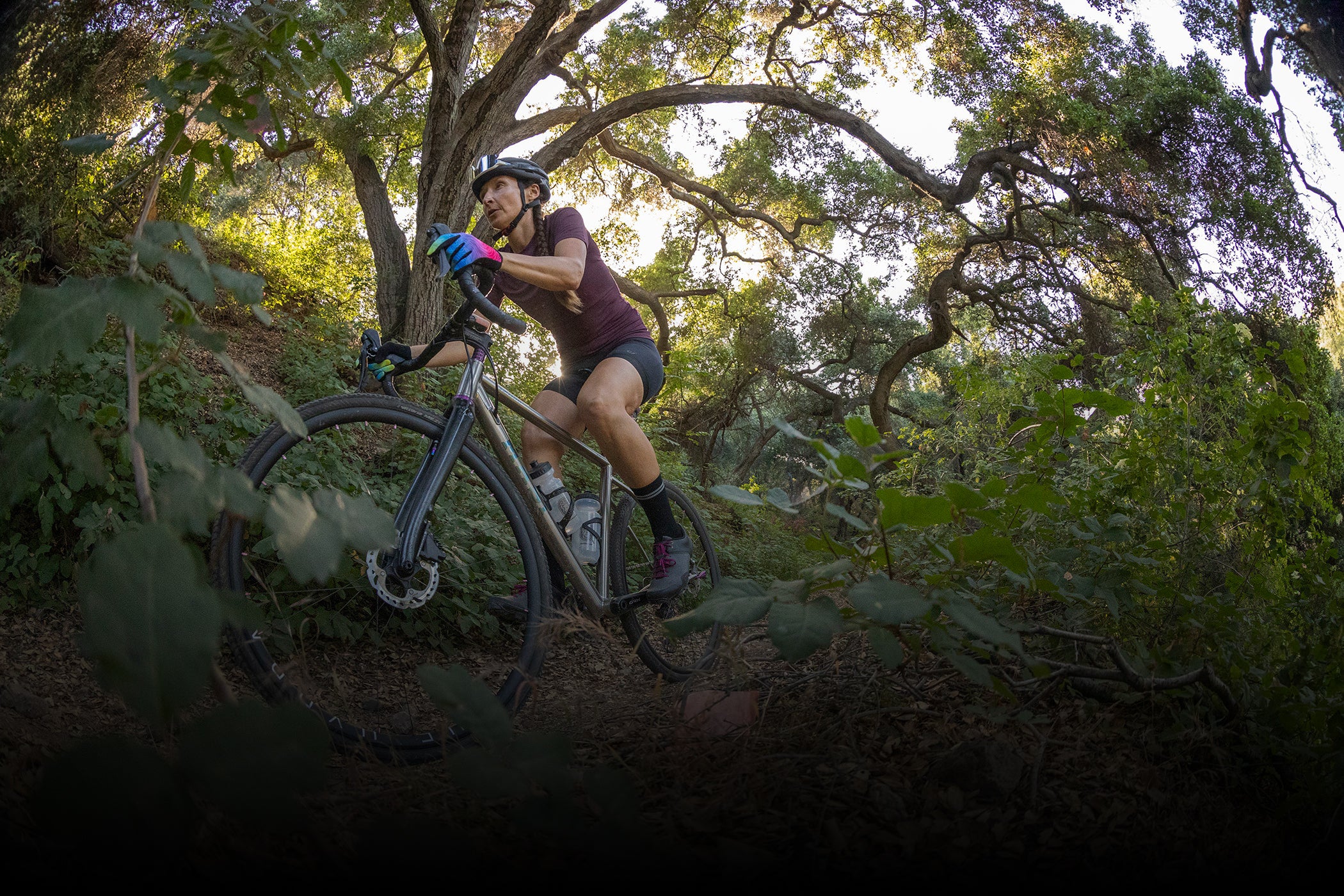A person wearing a helmet and gloves is cycling on a mountain bike through a dense, leafy forest trail. Sunlight filters through the trees, creating dappled shadows on the ground. The scene captures a sense of adventure and outdoor activity.
