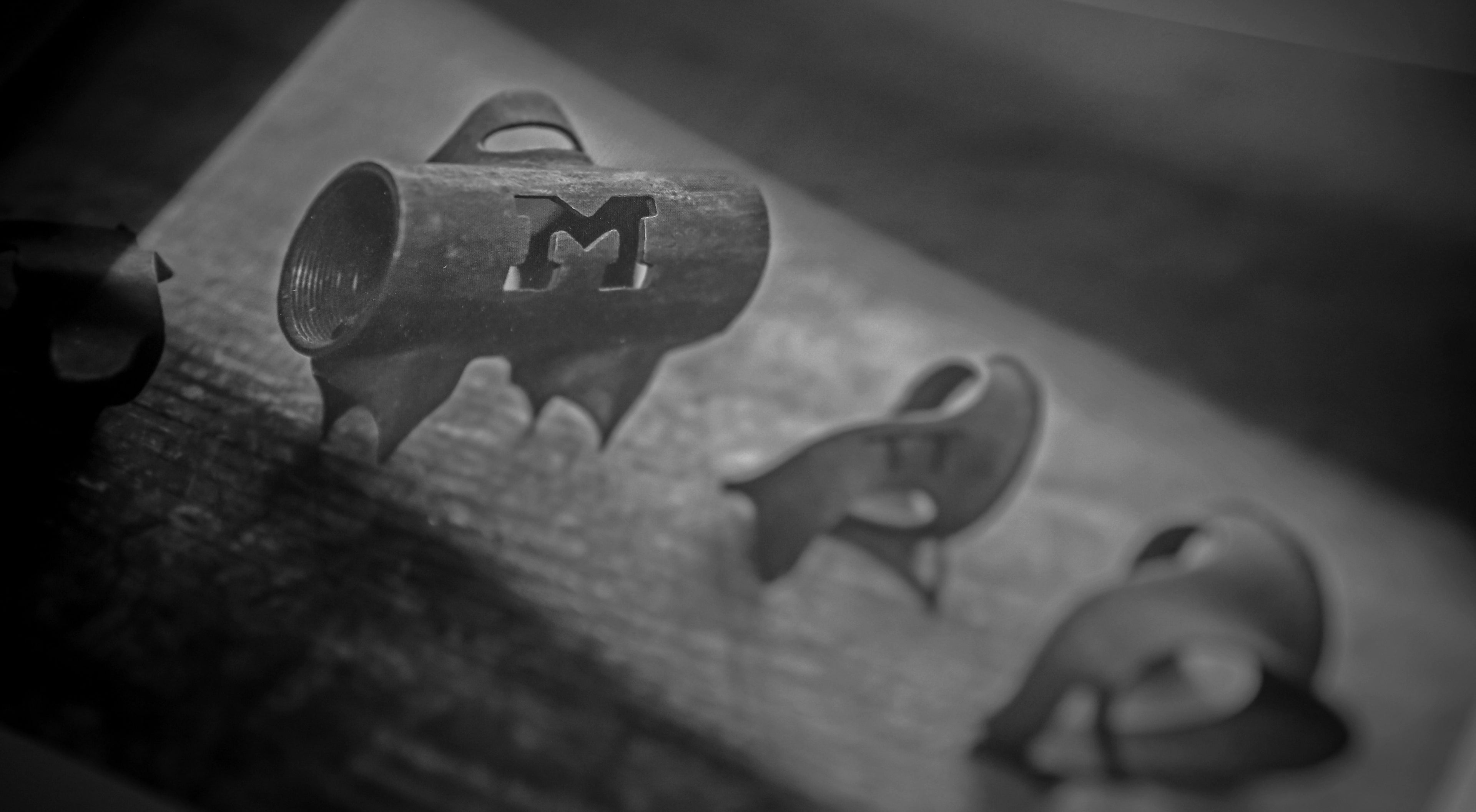 Close-up black and white photo showing three metal branding irons with handles. The central iron features a bold letter M, while the others appear partially visible. All are arranged on a wooden surface.