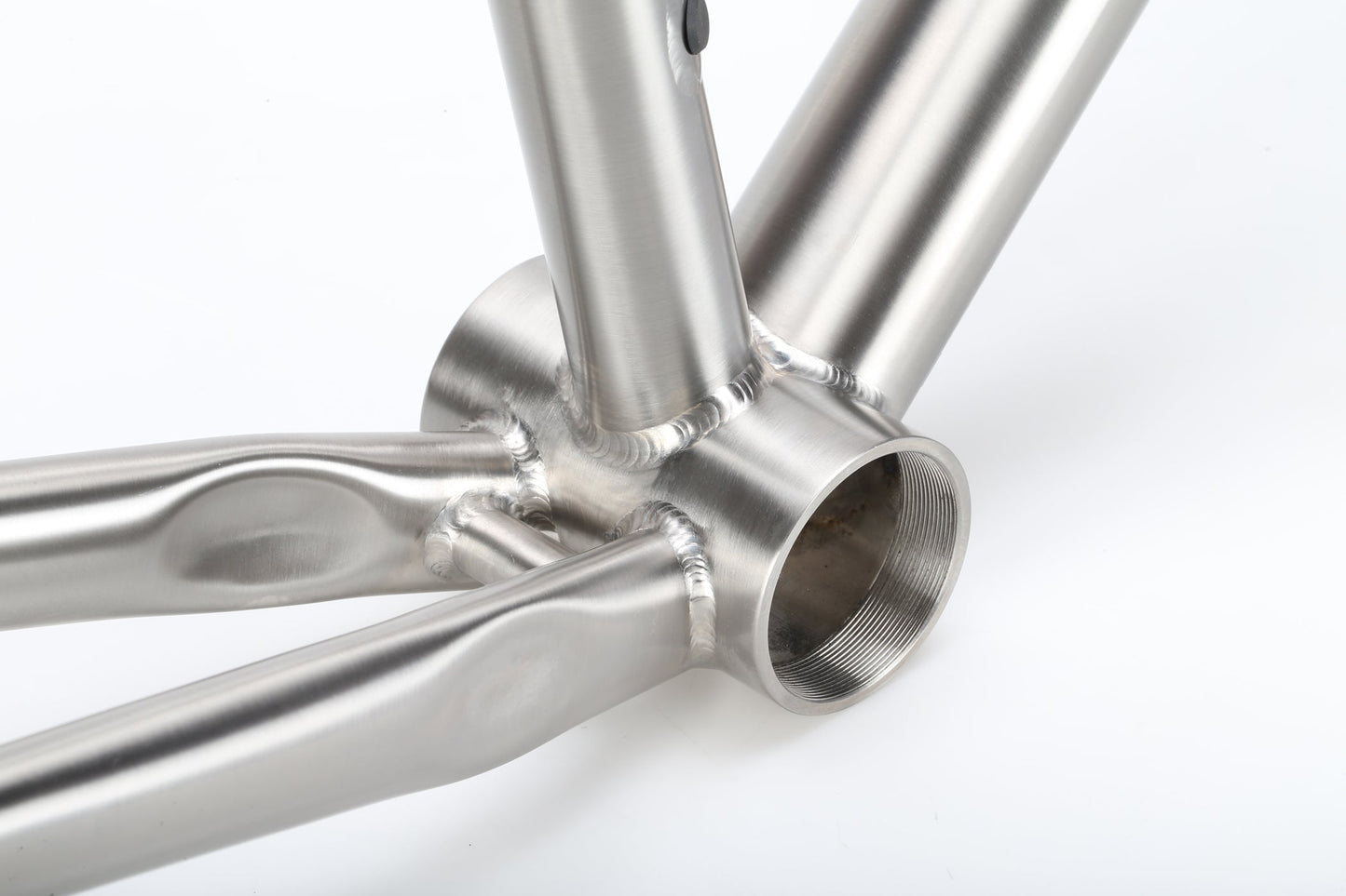 Close-up of a sleek TITANIO FRAMESET joint by Masi Bikes with smooth welds and polished finish, connecting the bottom bracket to chainstays and seat tube. The brushed texture and titanium race bike design highlight advanced craftsmanship with 32c tire clearance.