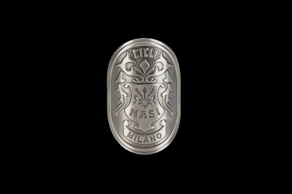Oval metal emblem with intricate engravings, featuring the words Cicli Masi Milano and decorative flourishes. The design is embossed, presenting a detailed and classic appearance against a black background.