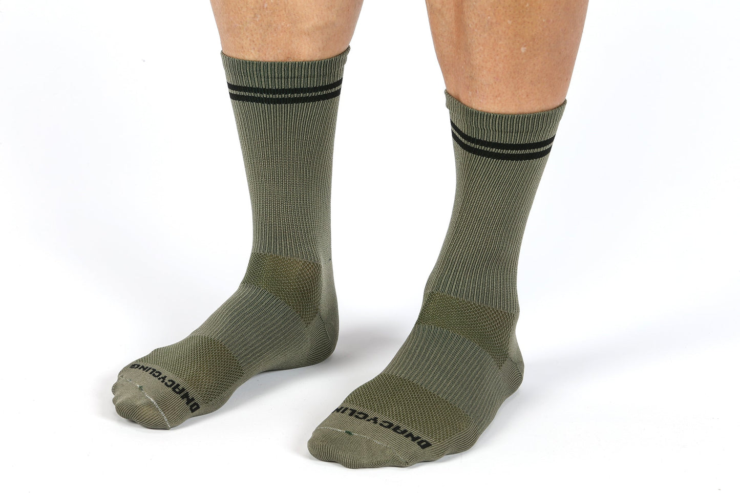 A pair of legs in olive green, sweat-wicking MASI DNA CYCLING SOCKS by Masi Bikes, featuring a black stripe near the top, labeled DONEGAL TRAIL on the toes. These performance socks have reinforced heels and toes and contrast beautifully against the minimalist white backdrop.
