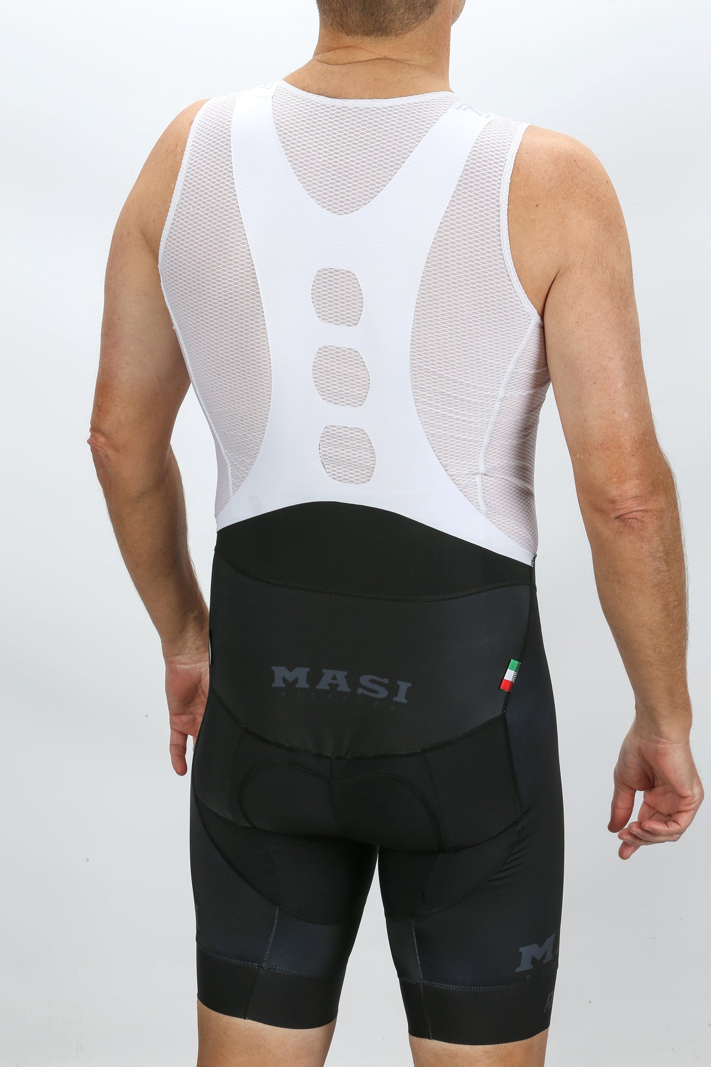 A person displays rider comfort from the back, wearing a white mesh sleeveless cycling jersey and black MASI DNA ELITE BIB by Masi Bikes. The shorts, ergonomically designed, have the MASI logo and an Italian flag detail on the side.