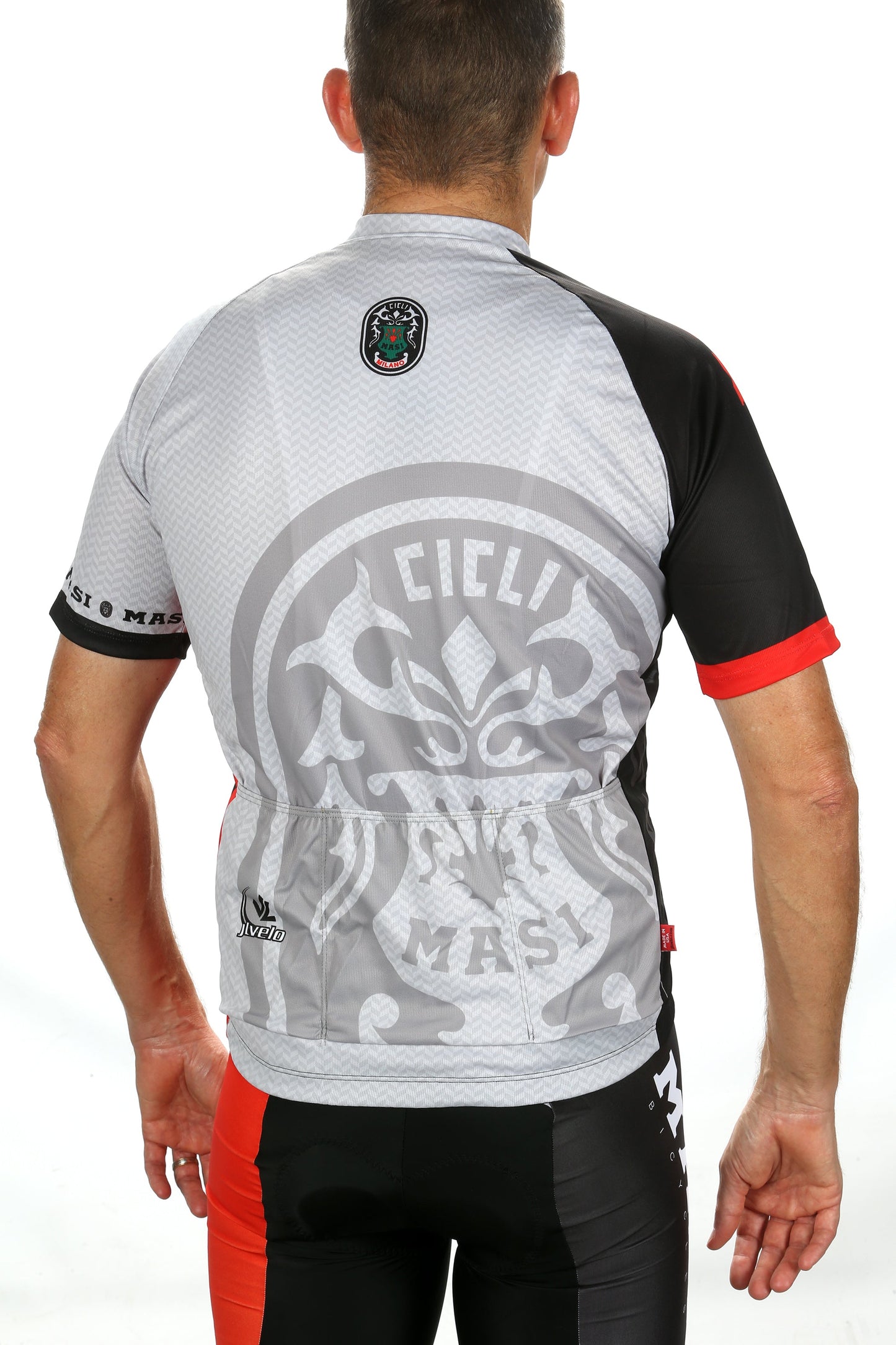 A cyclist wearing the MASI RGB JERSEY from Masi Bikes, featuring a large stylized Cicli Masi logo on the back in black, red, and gray colors, is facing away to showcase the jersey details.