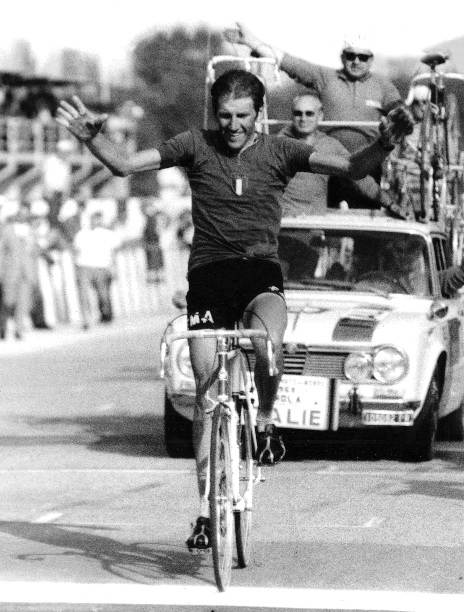A cyclist in a racing uniform crosses the finish line with arms raised in victory. A support car and cheering spectators are visible in the background.