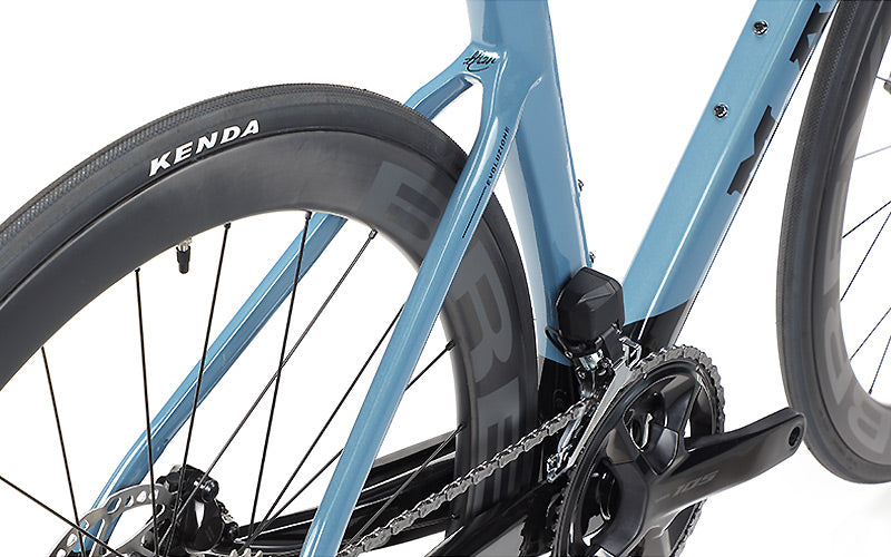 Close-up of a blue Masi Bikes EVOLUZIONE 1 Carbon bicycle featuring the KENDA branded rear tire, chain, and frame. The black chainring is mounted on the sleek, aerodynamic road design of the bike.