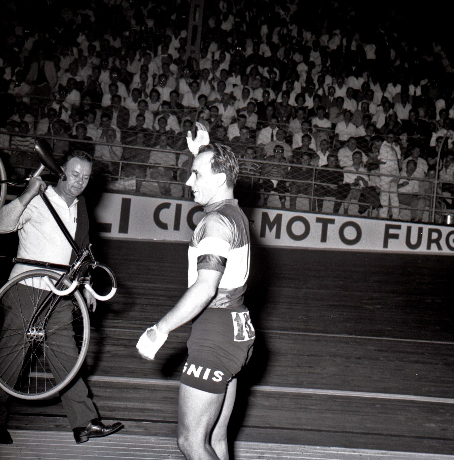 A cyclist wearing a numbered jersey walks on a velodrome track, holding a tire. Another person walks beside him, carrying a bicycle over their shoulder. The background is filled with a large crowd of spectators.