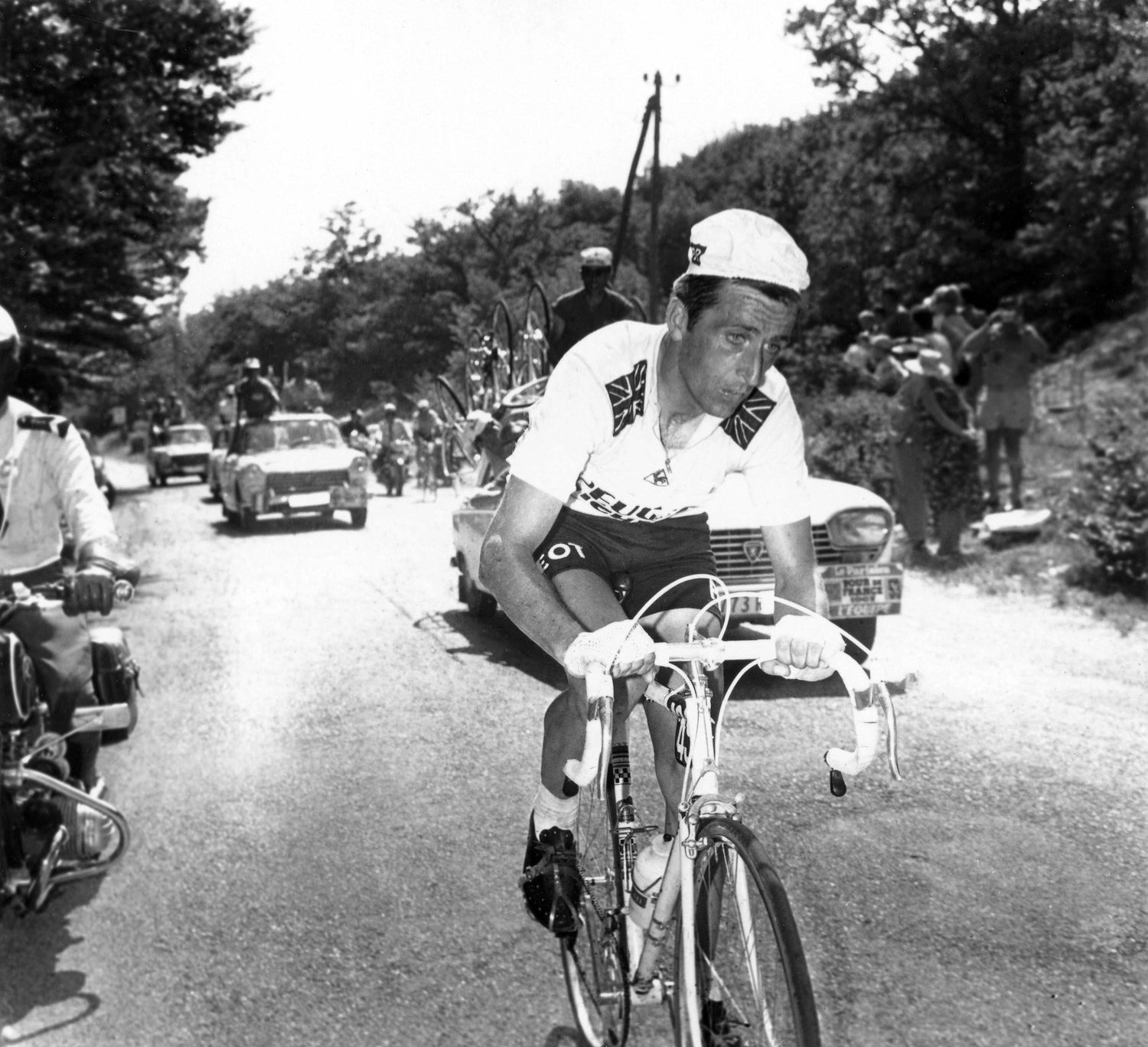 A cyclist in a racing outfit rides along a road flanked by trees, accompanied by a motorbike and followed by support cars. Spectators line the roadside, capturing the scene. The cyclist appears focused and determined.