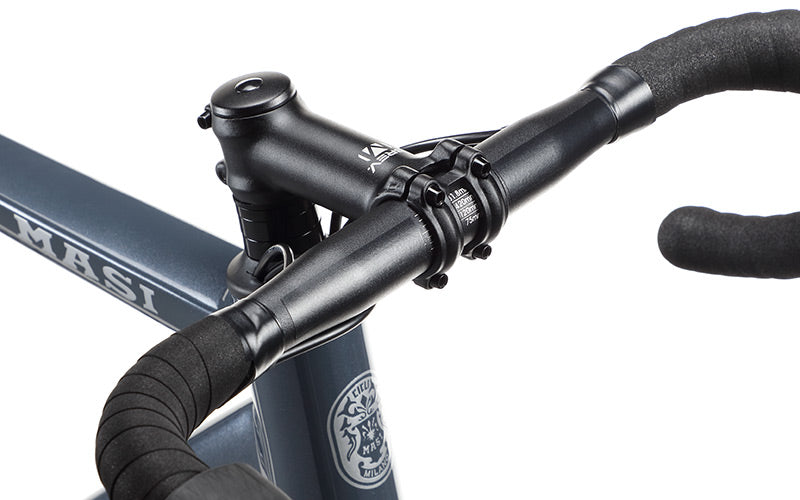 Close-up image of a bicycle handlebar and stem with black grips, set against a blue frame. The name Masi appears on the frame in white letters. The image showcases the bikes clean and modern design.