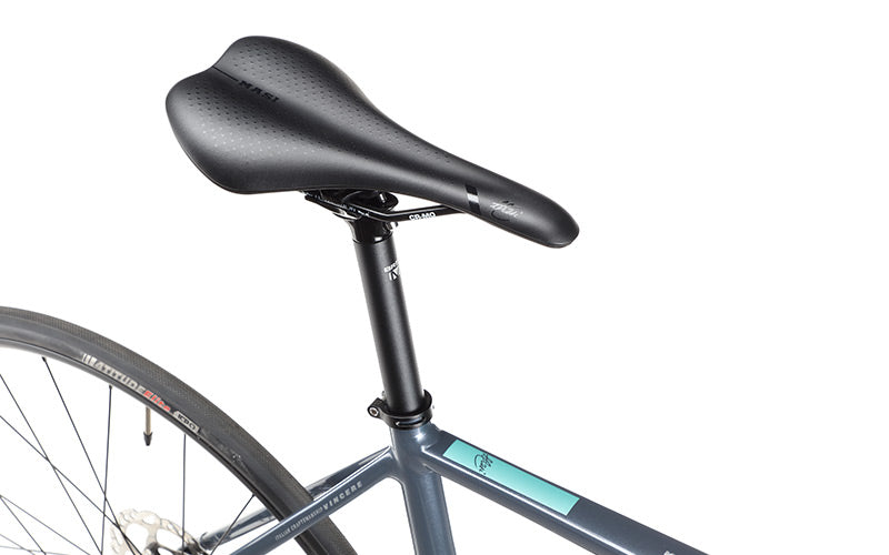 The image showcases a close-up of the VINCERE 3 bicycle seat, made by Masi Bikes, and a section of the alloy frame equipped with Shimano Tiagra. The black seat has small perforations and pairs with a dark gray frame accented in light blue, alongside the rear wheel on a white background.