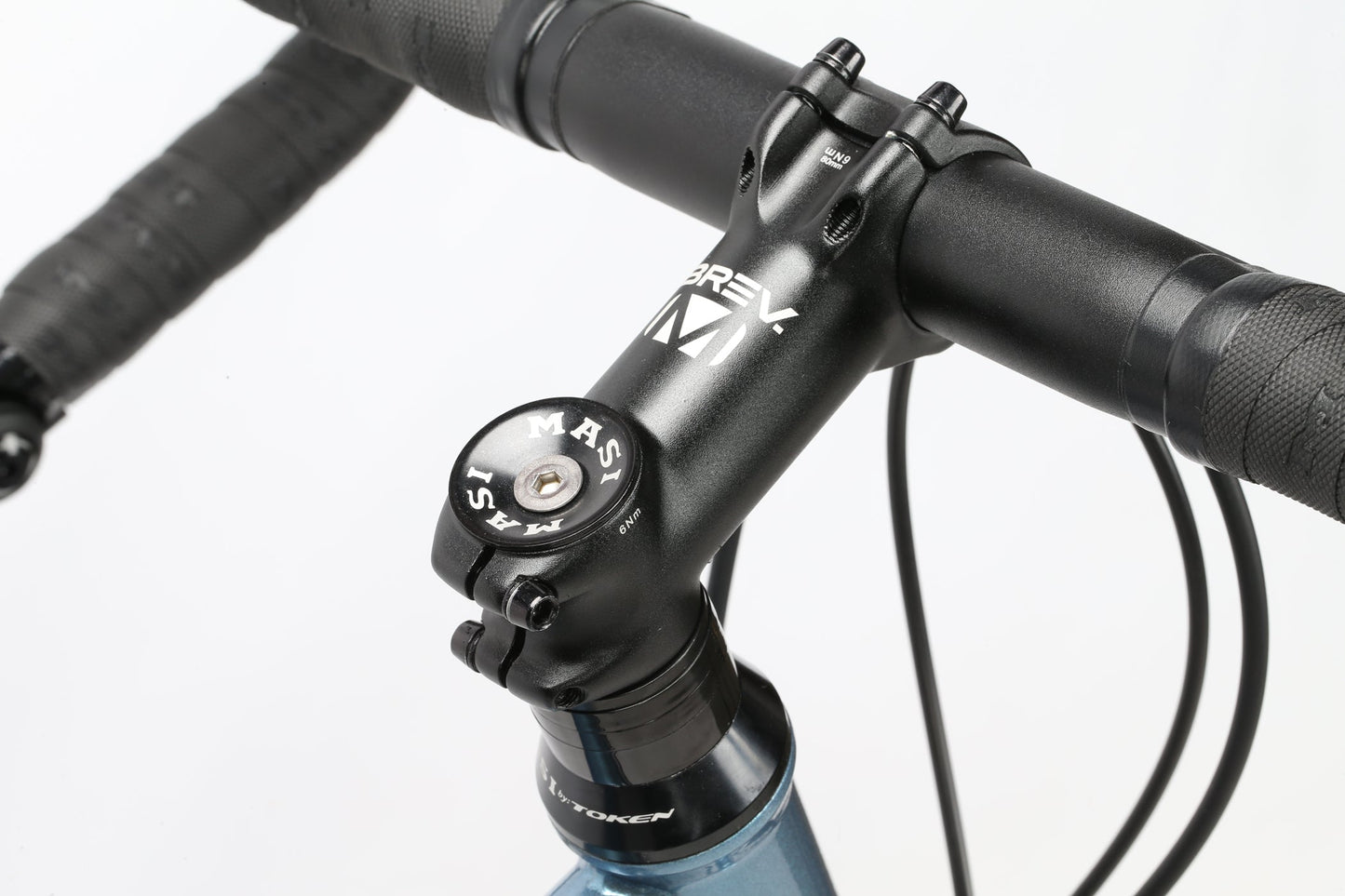 Close-up of a GIRAMONDO 700c touring bikes handlebar and stem assembly, with the metal stem labeled BREV and MASI. The textured black tape-wrapped handlebar, with visible cables, is ready for adventure on a crisp white background. Brand: Masi Bikes.