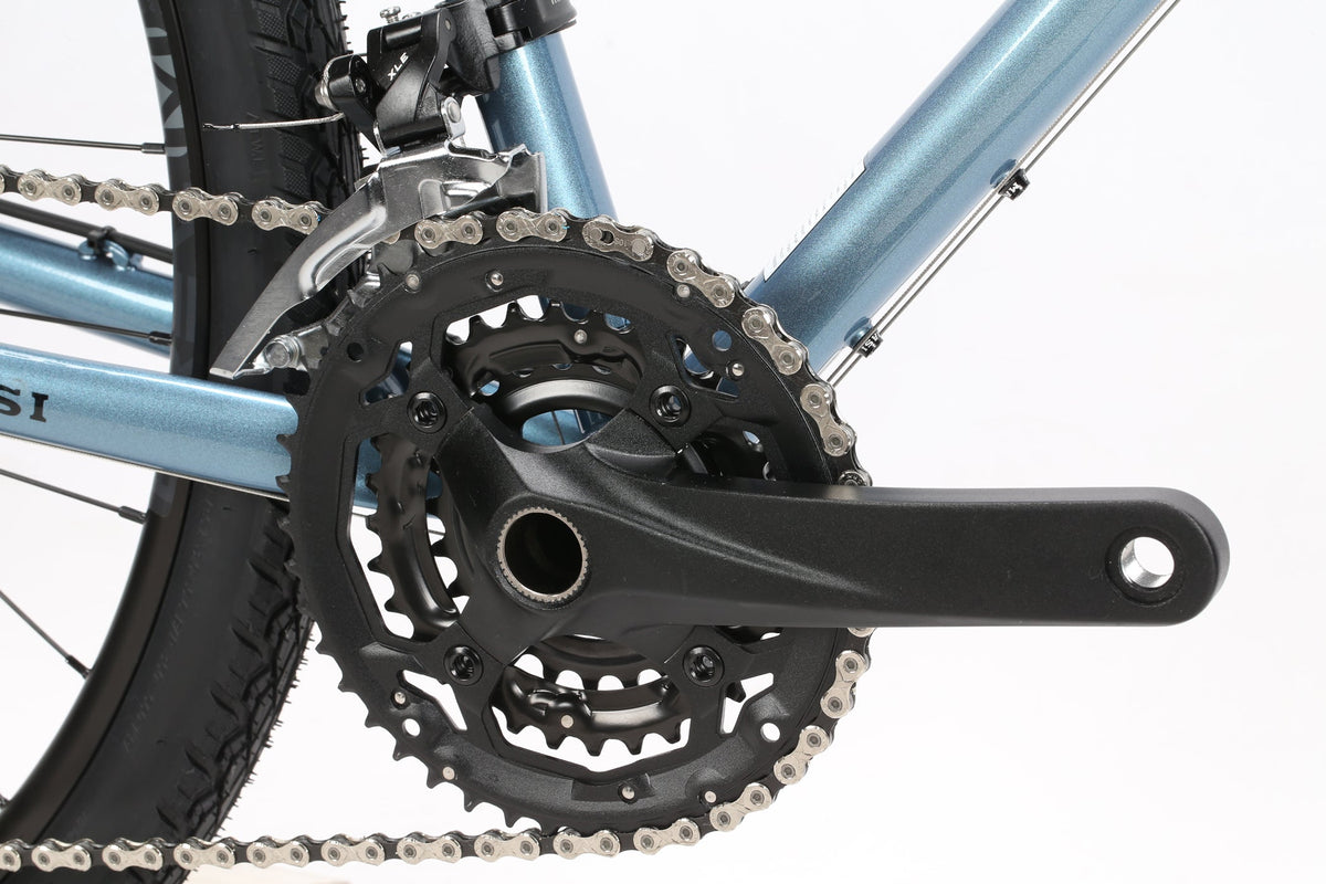Close-up of a Masi Bikes GIRAMONDO 700c touring bikes drivetrain, showcasing a black crankset with chainrings and a silver chain on a light blue frame. The focus is on the gear mechanism, capturing adventures essence with the tires tread pattern hinting at its Giramondo legacy.