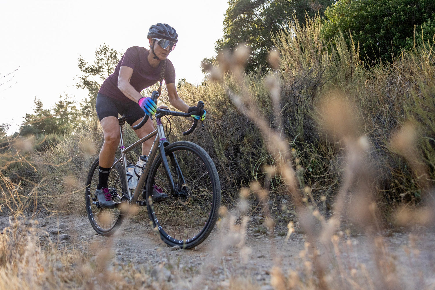 A cyclist wearing a helmet and sunglasses rides a gravel bike on a rough trail surrounded by dry vegetation. The sun casts a warm glow over the scene, highlighting the natural outdoor setting.