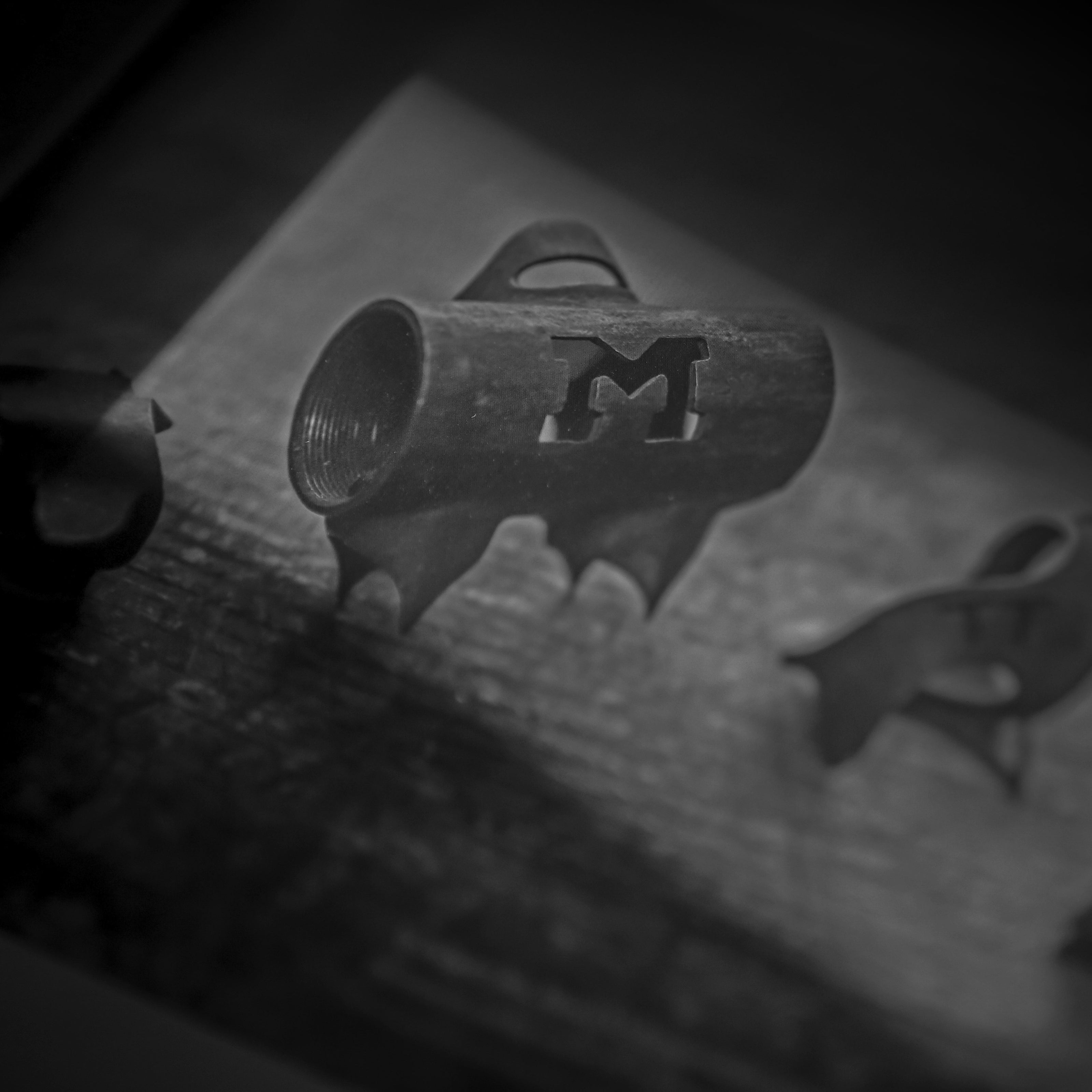 A black and white image of a metal object with a cutout of the letter M. The object is cylindrical and has claw-like extensions at the bottom. Its placed on a textured surface.