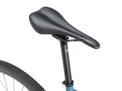 Close-up of the BRUNELLO 1s black saddle and seat post on a blue frame, with a knobby rear tire partly visible.