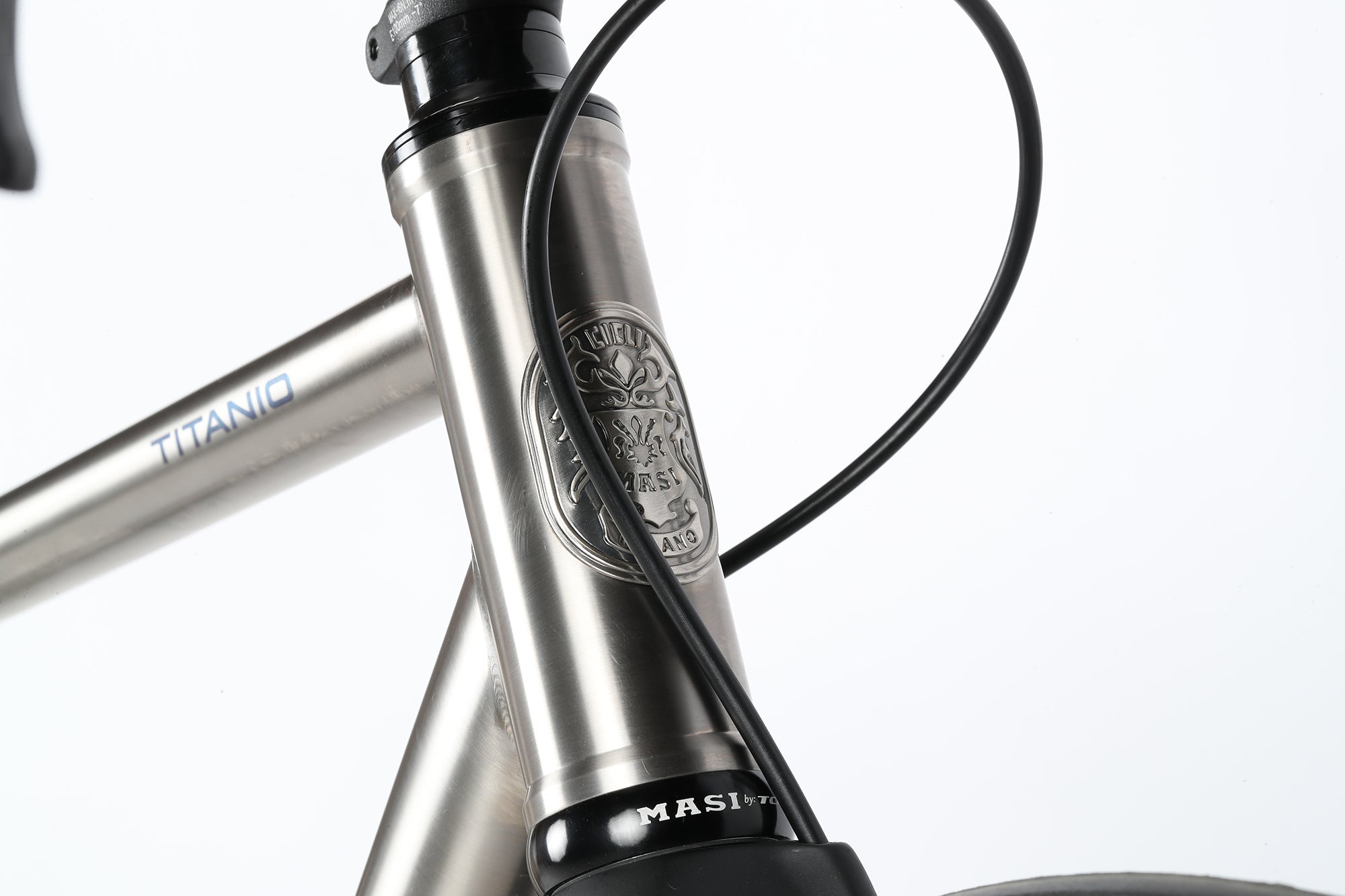 Close-up of a titanium bike frame with visible branding and intricate detailing on the head tube badge. Black cables are wrapped neatly along the frame, which displays a sleek, polished finish. The bike model name TITANIO is partially visible.