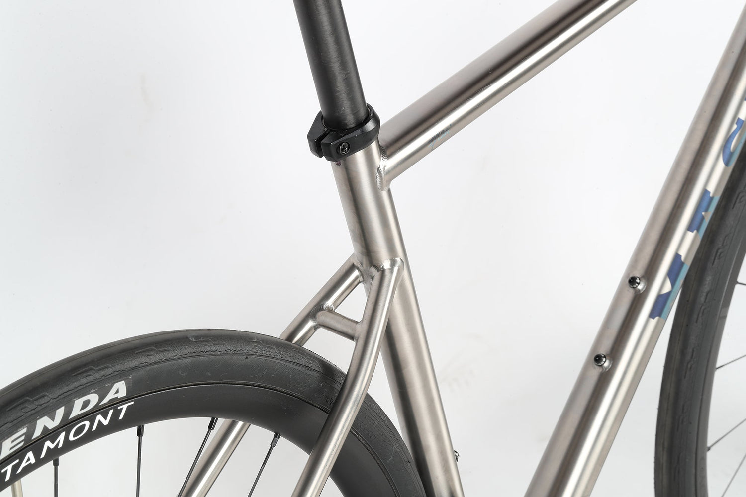 Close-up of a bicycle showing the rear section. The sleek metal frame has a polished finish, and the tire is branded KENDA. The image highlights the bikes clean design and sturdy construction against a plain background.